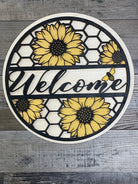 A circular wooden door hanger featuring a black and yellow honeycomb pattern with sunflower designs, and the word 'Welcome' in the center.