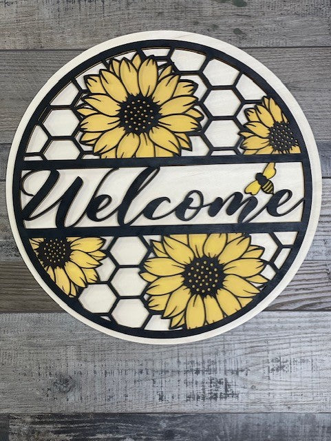 A circular wooden door hanger featuring a black and yellow honeycomb pattern with sunflower designs, and the word 'Welcome' in the center.